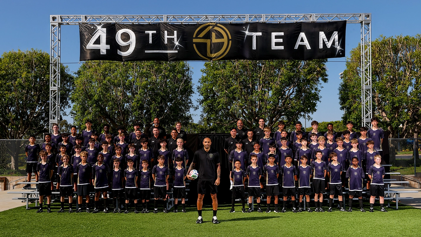 A 49th Team group photo of OMBC participants in navy kits gathered beneath a sign reading '49TH TEAM'