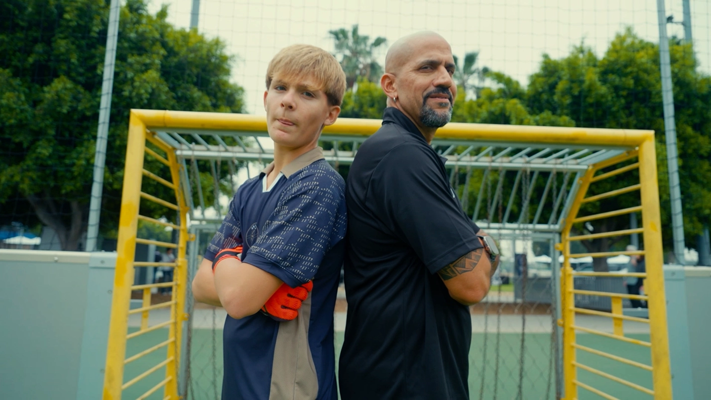 An OMBC participant and a coach standing back to back with arms folded in front of a goal on an outdoor pitch