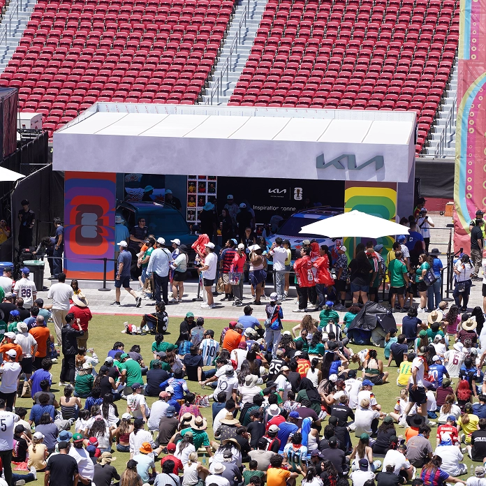 Fans seated on the grass around a Kia booth set up in front of the stadium stands