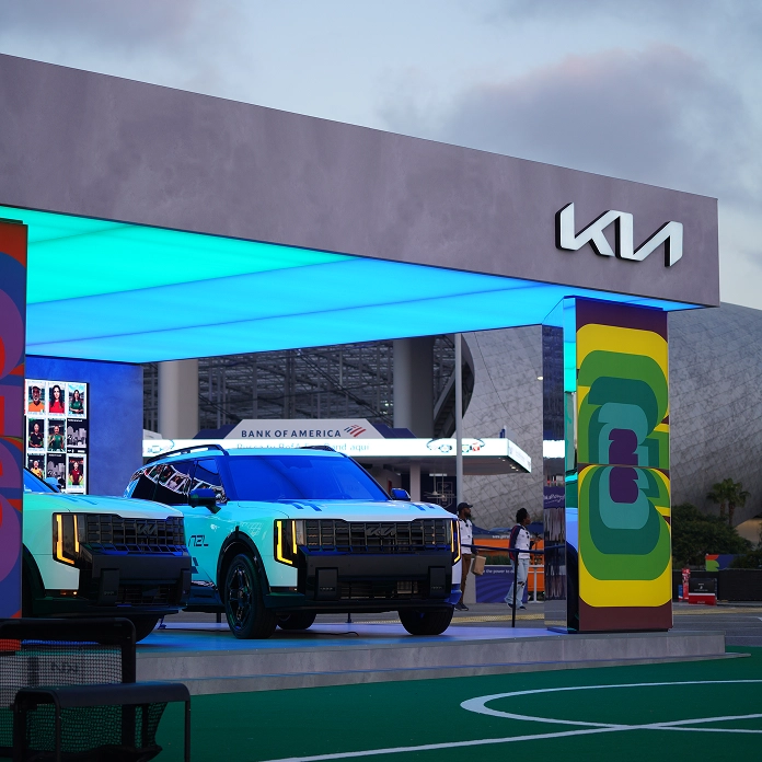 A Kia EV9 on display at a Kia booth outside a stadium, with a teal-lit canopy and a colourful patterned pillar