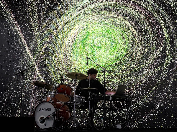 Live performance with drums and electronic equipment in front of particle-based visuals.