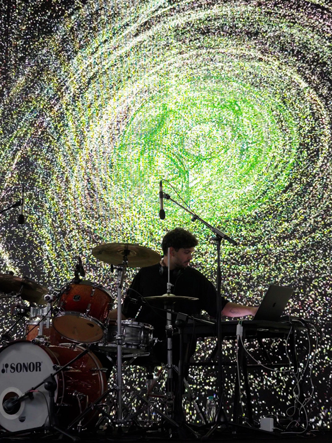Live performance with drums and electronic equipment in front of particle-based visuals.