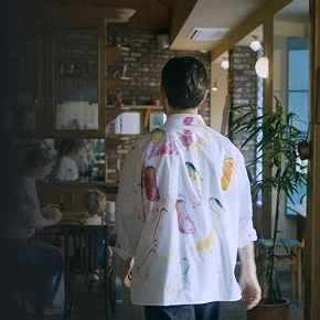 Back view of a person in a paint-stained shirt walking inside a café